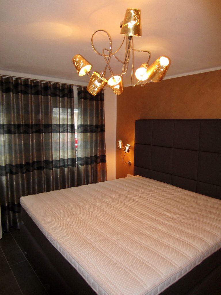 Die Ruheinsel in der Mitte des Schlafzimmers ist ein handgefertigtes Boxspringbett in ganz individuellen Maßen. Passend aufeinander abgestimmt dazu die Wandgestaltung und die Beleuchtung.