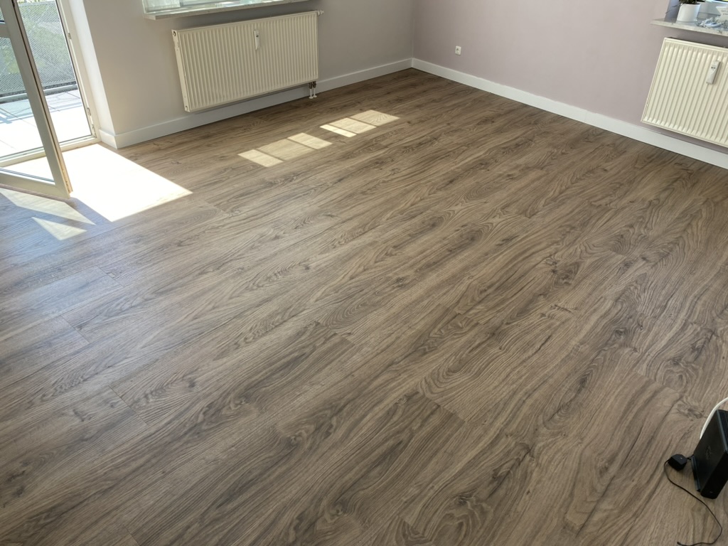 Fertiger Boden aus Vinyl in Landhausdielen-Optik im natürlichen Look und neuen schlanken weißen Sockelleisten, links die geöffnete Balkontür mit Sonnenlicht auf dem Boden
